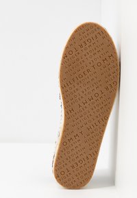 Semelle d'une chaussure avec « Tommy Hilfiger » embossé à plusieurs reprises en motif circulaire sur un matériau en caoutchouc marron clair.