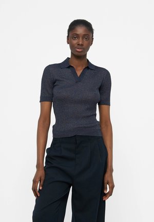 Γυναίκα με σκούρο δέρμα που φοράει στενό πλεκτό μπλουζάκι σκούρου μπλε χρώματος με κοντά μανίκια και φαρδιά μαύρη ψηλόμεση παντελόνα, στέκεται μπροστά σε λευκό φόντο.