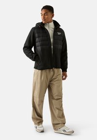 Czarna pikowana kurtka na zamek z kapturem, z czerwonymi akcentami, noszona na jasnoszarej bluzce. Beżowe spodnie o luźnym kroju i białe trampki.