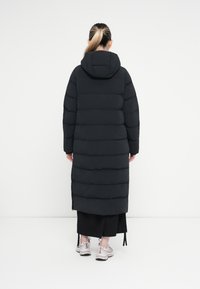 Melns pūkains mētis ar kapuci, ar horizontāliem quilted paneļiem un vidēja garuma dizainu. Nēsāts virs melnas svārku un pelēkiem sporta apaviem.