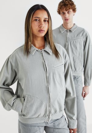 Deux modèles portent des vestes gris clair faites d'un tissu doux. Les vestes présentent une fermeture éclair à l'avant, un col et des poches ; un modèle se tient devant, l'autre derrière.