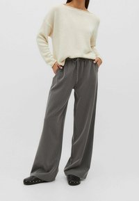 Helle beige Strickjacke mit weitem Ausschnitt, kombiniert mit grauen Streifen-Hosen mit weitem Bein, die einen elastischen Bund und eine Kordel haben. Schwarze Schuhe mit Nieten.