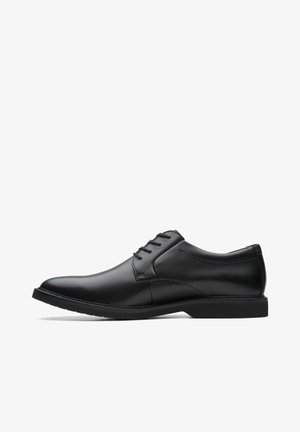 Chaussure habillée en cuir noir avec texture lisse, bout rond et cinq œillets pour les lacets. Semelle en caoutchouc noir à profil bas avec des détails de couture discrets.