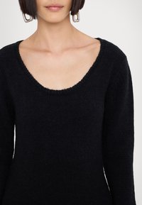 Femme portant un pull en maille noir avec un large col rond et de grandes boucles d'oreilles argentées rectangulaires.