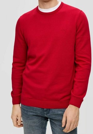 Pull en tricot rouge avec un col ras du cou, des poignets et un ourlet côtelés. Doté d'un logo discret sur la poitrine. Associé à un jean en denim bleu.