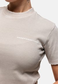 Maglietta beige a coste con scollatura a girocollo, bordi sfrangiati e testo bianco "GOODFOR NOTHING" sul lato sinistro. Tessuto morbido e testurizzato.