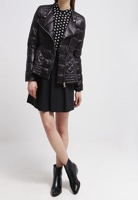 Sort pufferjakke med en diagonal lynlås, kombineret med en sort og hvid prikket top og en sort nederdel, suppleret med sorte ankelstøvler.