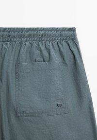 Pantaloni corti con cintura elastica in tessuto grigio chiaro, caratterizzati da una tasca posteriore singola con pattina e un dettaglio in metallo vicino alla tasca.