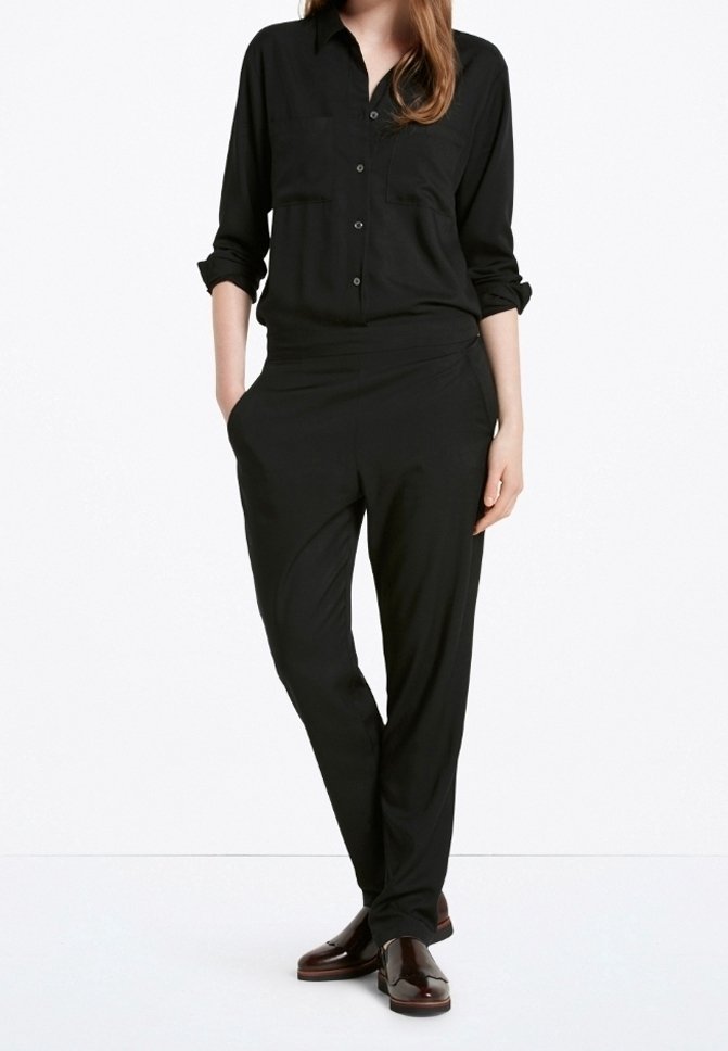 Femme portant une chemise noire boutonnée avec manches retroussées, un pantalon noir fuselé avec poches, et des mocassins marron foncé.