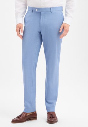 Mann trägt hellblaue Anzughose, weißes Hemd und braune Slipper mit Quasten, steht mit entspannten Händen an den Seiten.