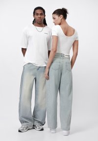 Två personer i avslappnade outfits: en vit oversized t-shirt och ljusblå vida jeans med en blekt gradient. Kvinnans topp har en bred halsringning.