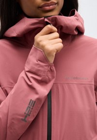 Άτομο που κλείνει το φερμουάρ σε ροζ μπουφάν Peak Performance με ψηλό γιακά και εκτυπωμένο λογότυπο στο μανίκι.