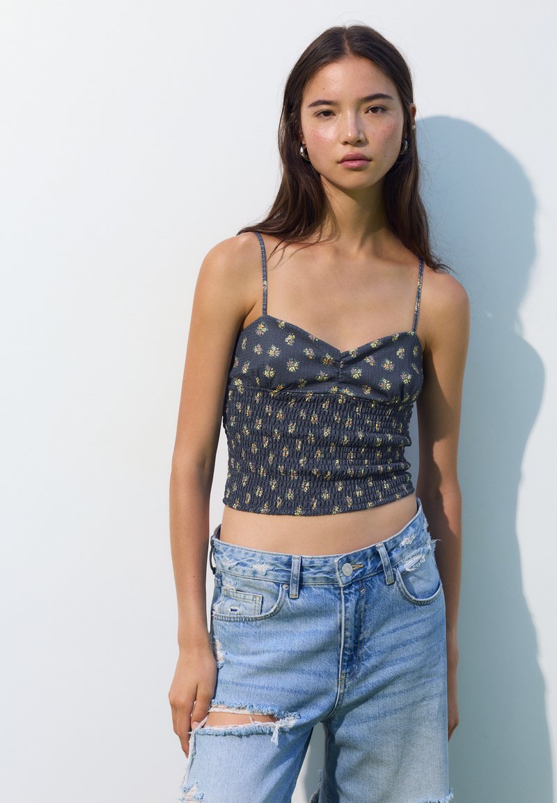 Top corto blu scuro con motivo floreale colorato, texture arricciata e spalline sottili, abbinato a jeans chiari strappati.