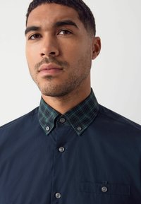 Marine blouse met een geruite groene kraag. Glad materiaal, twee knopen bij de kraag en een borstzak met een accent van bijpassende stof.