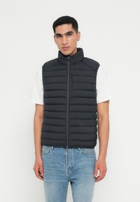 Sort puffervest med vertikal kviltet design, høj krave og lynlås, parret med en hvid skjorte og lyseblå jeans.