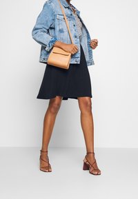 Džínová bunda, černé šaty s rozšířeným lemem, béžová přes rameno taška a vínové sandály na podpatku. Jednoduchý design, kontrastní barvy, různé textury.