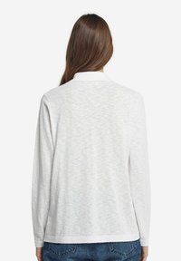 Vit långärmad polo-shirt med krage, med ett subtilt strukturerat mönster och rak nederkant. Bakifrån visas en enkel design.