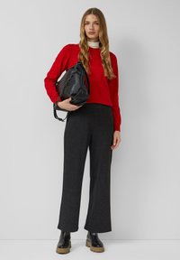 Roter Strickpullover, schwarze lässige Handtasche mit Kettenriemen, graue weit geschnittene Hose und schwarze Stiefel mit klobigem Absatz. Schlichter Hintergrund.