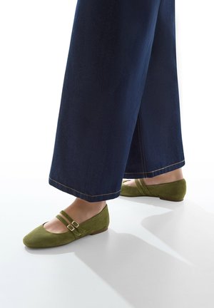 Ballerine in suede verde con due cinturini sottili e fibbie dorate. Abbinate a pantaloni in denim blu scuro a gamba larga. Texture liscia, design con punta arrotondata.