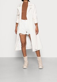 Kobieta w długim białym płaszczu, brązowym prążkowanym golfie, białych podartych szortach i kremowych botkach sznurowanych na kostkę.