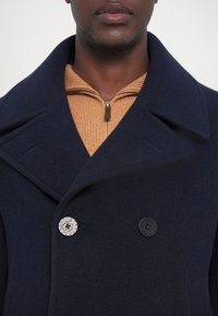 Mornarski kaput od vune s dodatkom, s velikim reverima, srebrnim i crnim dugmadi te bež džemperom s lažnim ovratnikom ispod. Vidljiva teksturirana tkanina.