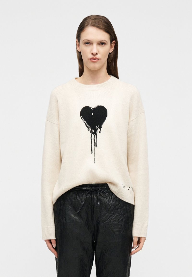 Γυναίκα που φοράει κρεμ πουλόβερ με μαύρη καρδιά που στάζει και μαύρο παντελόνι με κορδόνι, στέκεται μπροστά σε απλό λευκό φόντο.