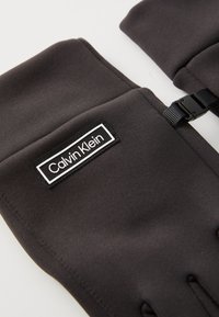 Schwarze Stoffhandschuhe mit einem Calvin Klein-Logo, schlankem Design und einer seitlichen Schnalle zur Anpassung. Glatte Textur mit maßgeschneiderten Fingerabschnitten.