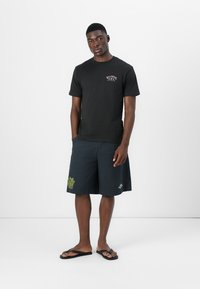 Svart bomulls t-skjorte med logo på fronten, kombinert med marineblå shorts med grønt bladdesign. Modellen har på seg svarte flip-flops. Minimalistisk bakgrunn.