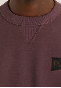 Felpa color marsala con colletto a coste, dettaglio di cucitura a V, e una patche nera con un design arancione sul lato inferiore.