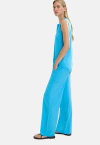 Niebeżowy top bez rękawów i dopasowane szerokie spodnie wykonane z gładkiego materiału. Modelka stoi z boku, widoczne kieszenie, w płaskich sandałach.