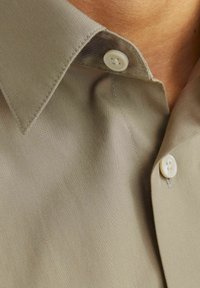 Lichtbeige shirt met een puntcollar, twee witte knopen en een soepele stoftextuur met subtiele stiksels.