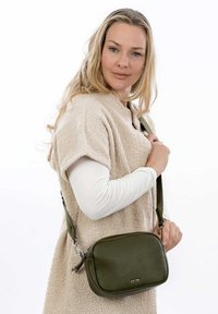 Borsa a tracolla in pelle verde con finitura texturizzata, chiusura con zip e tracolla regolabile, indossata su un indumento di colore neutro.