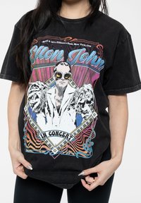 Black oversized graphic t-shirt featuring Elton John illustration with colorful accents, text, and a retro design. Soft cotton fabric.