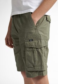 Shorts cargo vert olive en tissu léger. Comprend des poches latérales et des détails en rabat, avec une petite étiquette de marque sur le côté gauche.