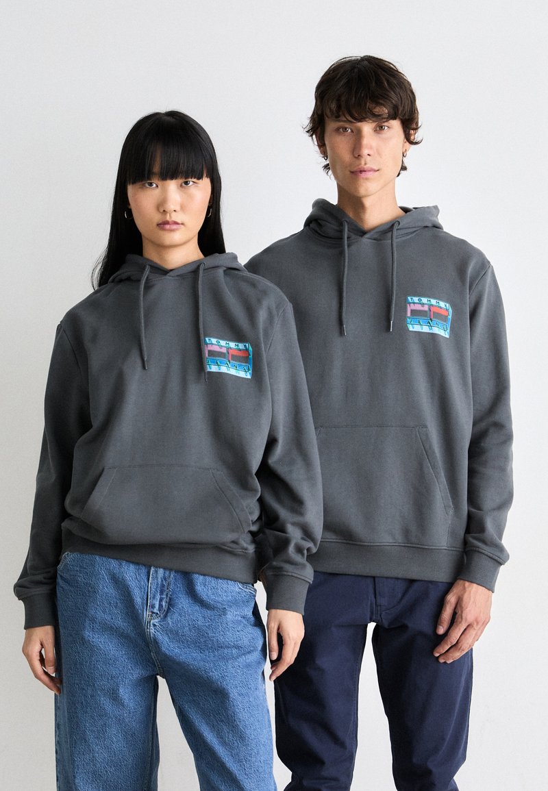 Grijze hoodies met een voorkangoeroezak, voorzien van een kleurrijke grafische patch op de borst. Draag deze met blauwe en marineblauwe broeken door twee modellen.