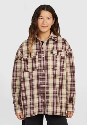 Jong persoon met lang haar draagt een oversized beige en bruine geruite blouse met borstzakken, staand tegen een effen achtergrond.