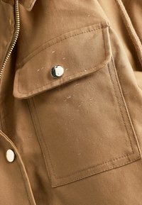 Gros plan sur la poche d'une veste en tissu marron avec un bouton-pression argenté et une fermeture éclair visible sur le côté.