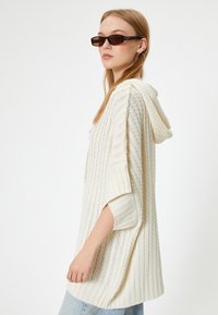 Sudadera de punto en color crema con textura acanalada y diseño estampado. Cuenta con mangas amplias y una capucha ajustable. Se lleva con gafas de sol oscuras.