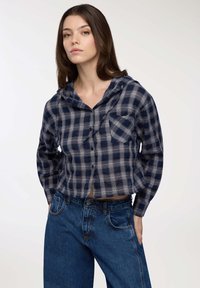 Giovane donna con lunghi capelli castani che indossa una camicia a maniche lunghe con cappuccio a quadri blu e grigi e jeans blu a vita alta, con le mani nelle tasche.
