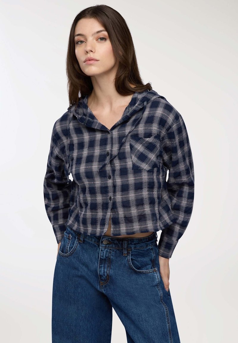 Giovane donna con lunghi capelli castani che indossa una camicia a maniche lunghe con cappuccio a quadri blu e grigi e jeans blu a vita alta, con le mani nelle tasche.