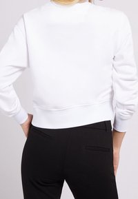 Felpa corta bianca con polsini e orlo a costine; texture liscia. Indossata con pantaloni neri con tasche posteriori sottili.