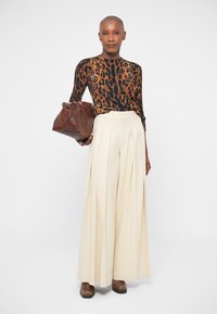 Braunes und schwarzes Leopardenmuster Langarmoberteil, hochtaillierte plissierte Cremhosen, braune Ledertasche, braune Schuhe. Schlichter Hintergrund.