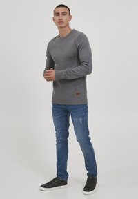 Grijze gebreide trui met geribde manchet en zoom, gecombineerd met blauwe jeans en zwarte sneakers. Beschikt over een klein bruin leren logo-patch op de trui.