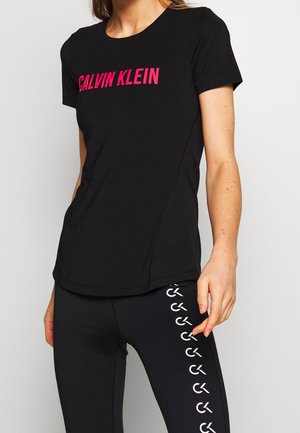 Femme portant un t-shirt noir Calvin Klein avec texte rose et des leggings noirs avec un motif vertical blanc du logo CK sur le côté.