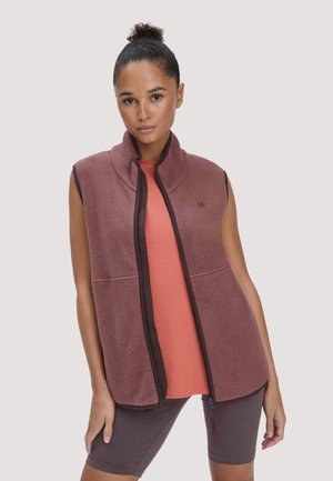 Frau mit lockigem Dutt trägt eine ärmellose mauvefarbene Fleeceweste über einem korallfarbenen Shirt und dunkel mauvefarbenen Radsportshorts, steht vor einem einfarbigen Hintergrund.