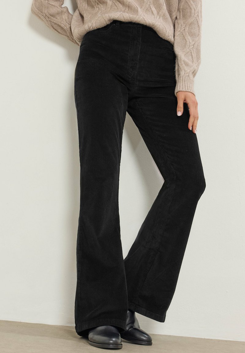 Zwarte fluwelen flare pants met verticale ribbels, hoge taille en een lichte glans. Gepaarde met zwarte schoenen en een gestructureerde beige gebreide trui.
