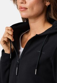 Zwarte zip-up hoodie met een gladde textuur, verstelbare trekkoorden en subtiele logo-details op de borst. Rits heeft accenten van zilverkleurig hardware.