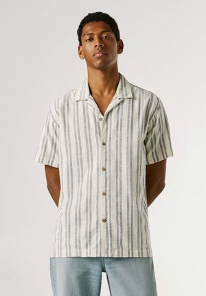 Giovane uomo che indossa una camicia a maniche corte con bottoni e righe verticali blu e jeans azzurri, in piedi con le mani dietro la schiena.