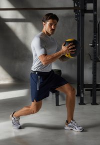Atletický muž wearing fitted šedé tričko s krátkym rukávom a námornícke šortky, drží čiernožltú medicínsku loptu, na pozadí telocvične.