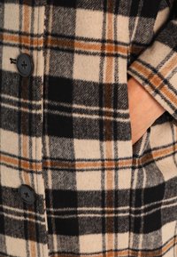 Takkivillaneule, jossa on beige, musta ja oranssi ruutukuosi, mustat napit ja sivutaskut lisätoiminnallisuutta varten.